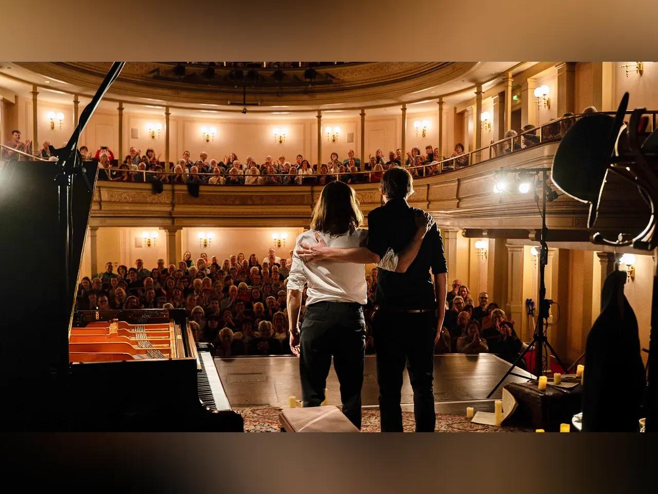 Das Klavierduo von hinten auf einer Bühne mit einem schicken Saal und Publikum im Hintergrund