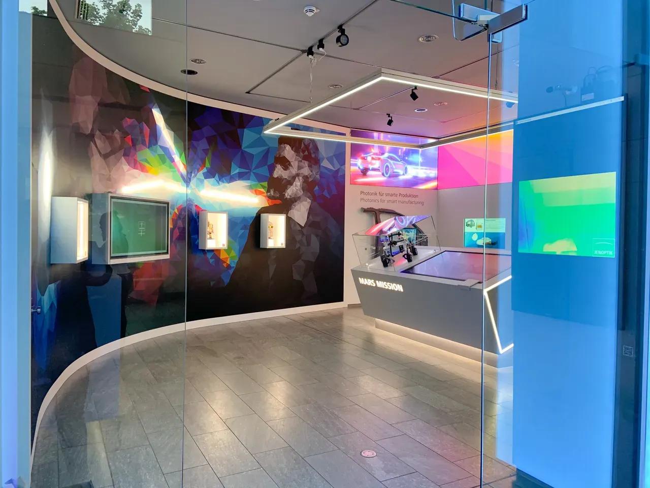 Ausstellungsbereich der JENOPTIK AG mit beleuchteten Vitrinen, Produkten und einem bunten Wandgemälde mit dem abstrahierten Portrait von Carl Zeiss