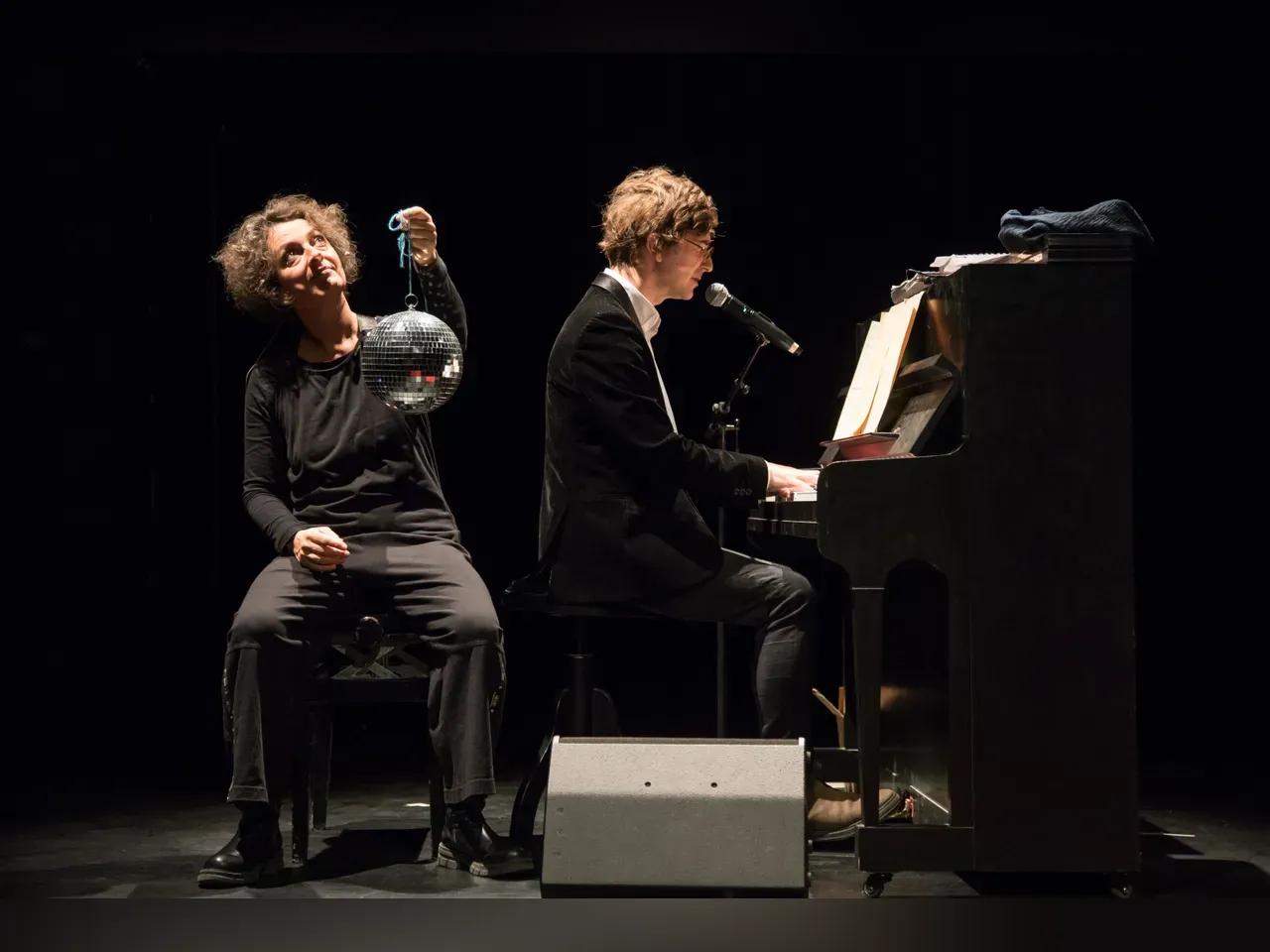 Antje Horn auf einem Stuhl mit einer Discokugel in der Hand und Stefan Nagler singend am Klavier