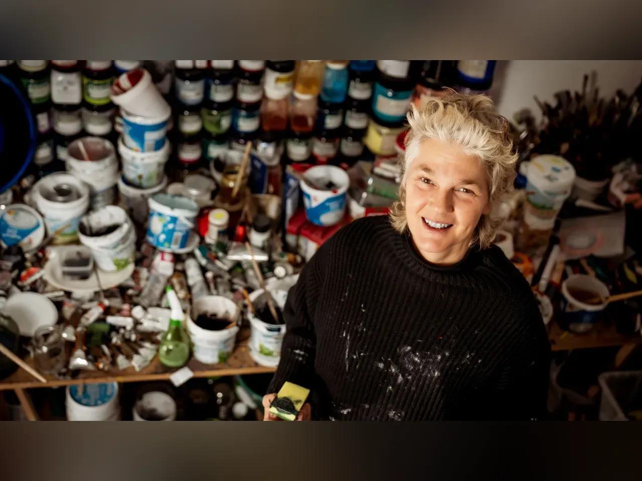 Frau mit relativ Kurzen Haaren steht in einem Atelier zum Kamera gerichtet und lächelt