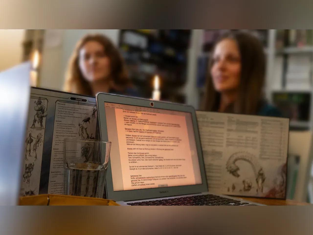 Es ist ein Laptop mit Text zu sehen, dahinter ein Pen und Paper Schichtschutz mit Regeln daruf. Im Hintergrund sieht man Kerzen und zwei Spielerinnen. 