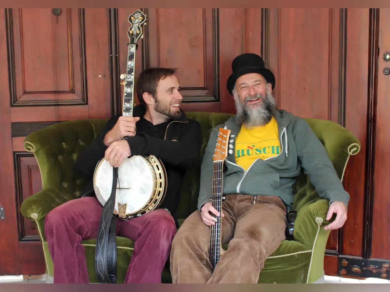 Die zwei Musiker "Doc Taylor" mit Banjo und Gitarre sitzen lachend auf einem Sofa