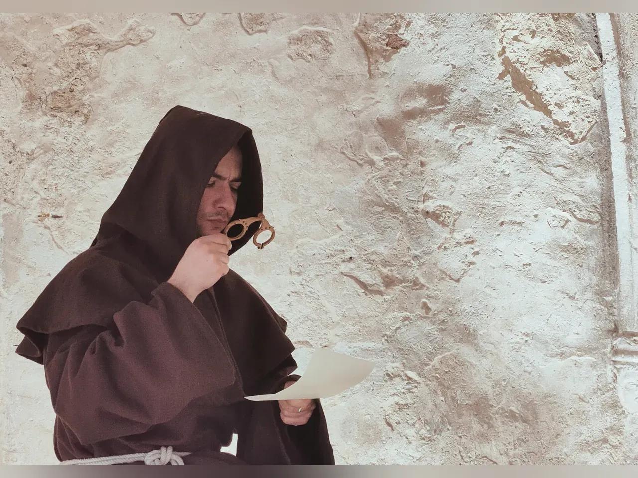 Mönch in brauner Kutte mit Brillengestell und Schriftstück in der Hand, sitzend  in einem Natursteingewölbe