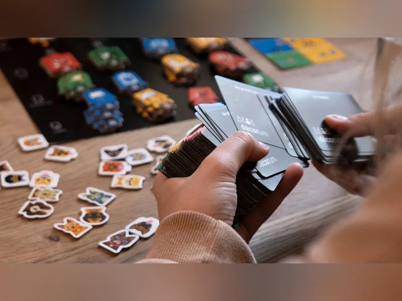 Eine Person schaut Spielkarten in ihrer Hand an. Auf dem Tisch im Hintergrund ist ein Spielbrett aufgebaut. 